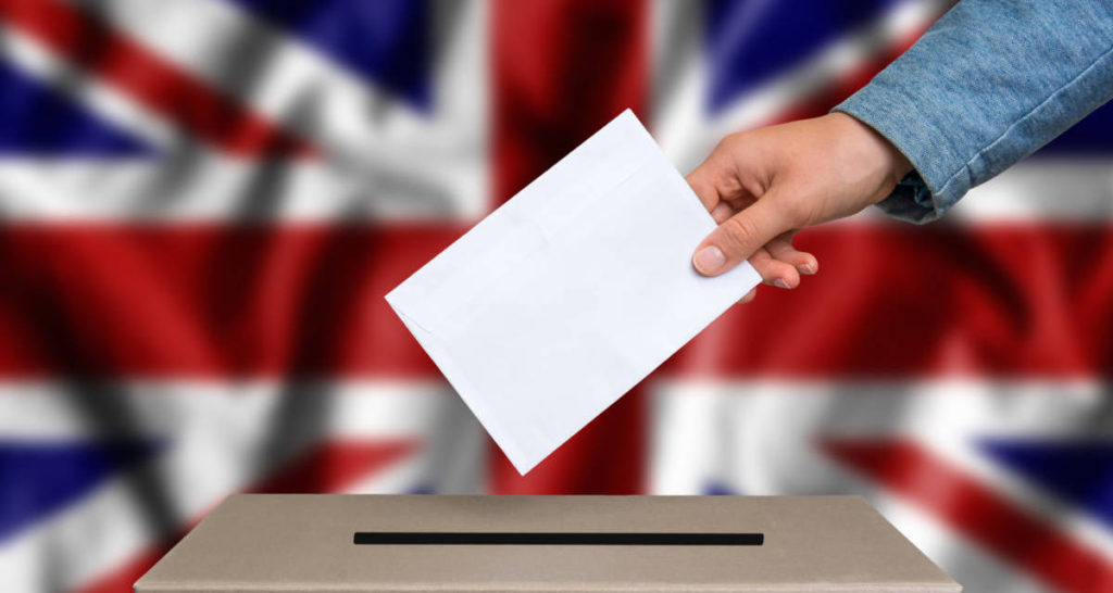 Election in Great Britain. The hand of woman putting her vote in the ballot box. British flag on background.